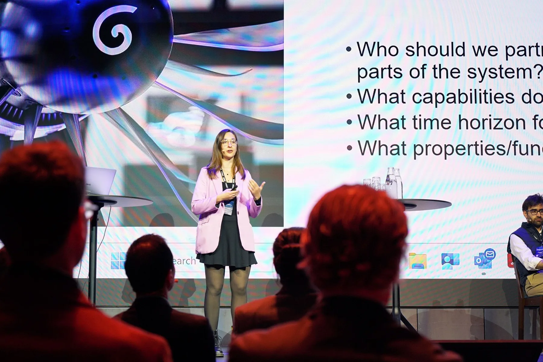 A group member presenting research on a conference stage, in front of a slide showing an aircraft engine