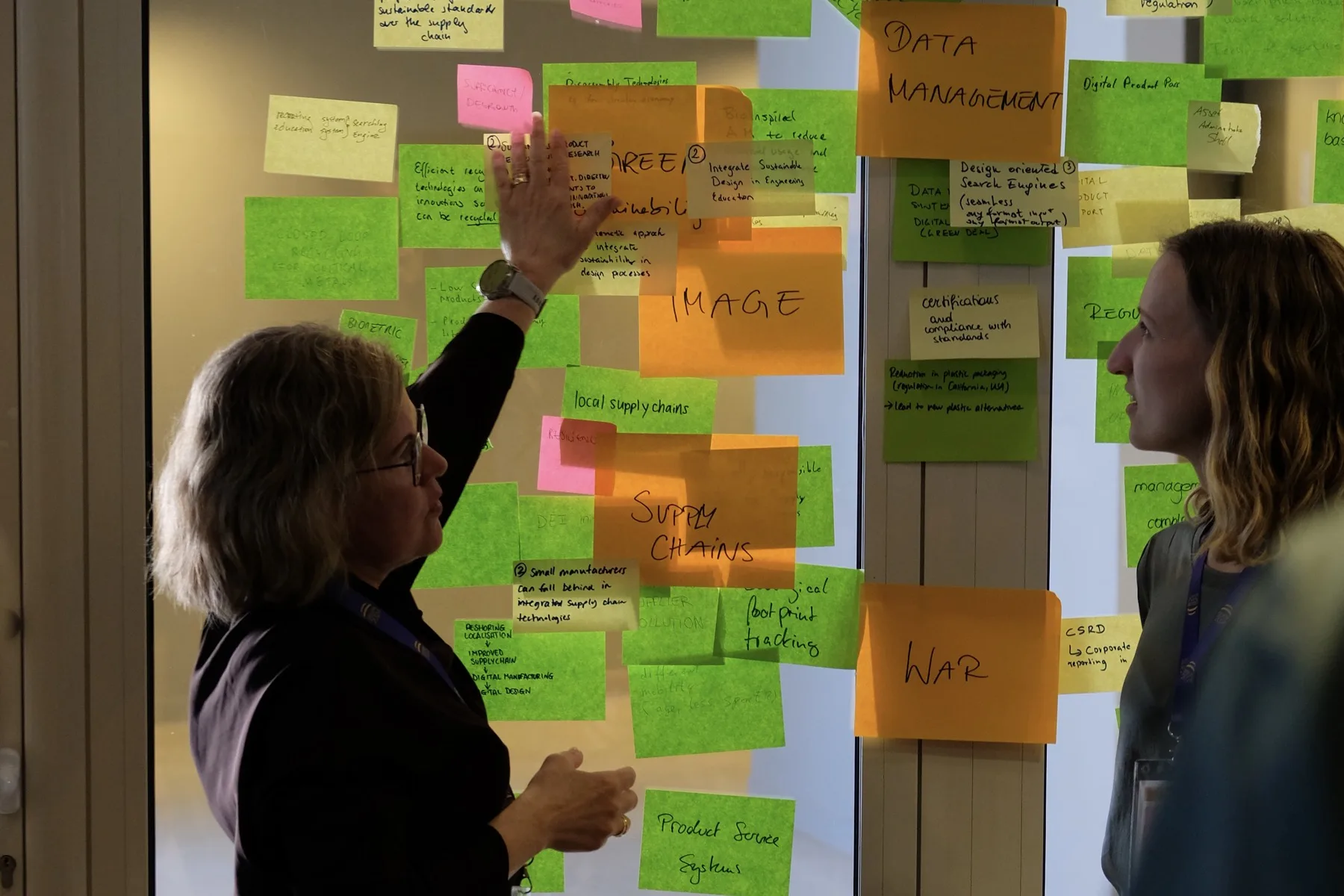 Two researchers clustering sticky notes on a glass wall during a project workshop
